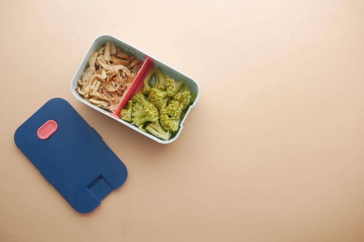 Kotak bekal berisi mi dan brokoli dengan sekat merah di atas meja berwarna krem, tutup biru dengan aksen merah di sampingnya.