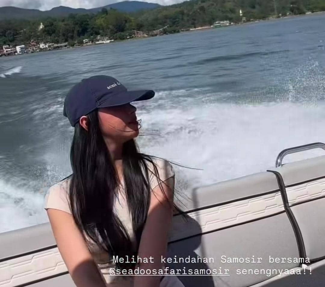 Seorang perempuan berambut panjang mengenakan topi biru duduk di perahu motor di atas Danau Toba dengan latar Pulau Samosir yang hijau.