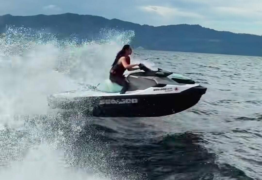 Seorang perempuan mengendarai jet ski di atas air dengan latar pegunungan dan langit cerah di Pulau Samosir.