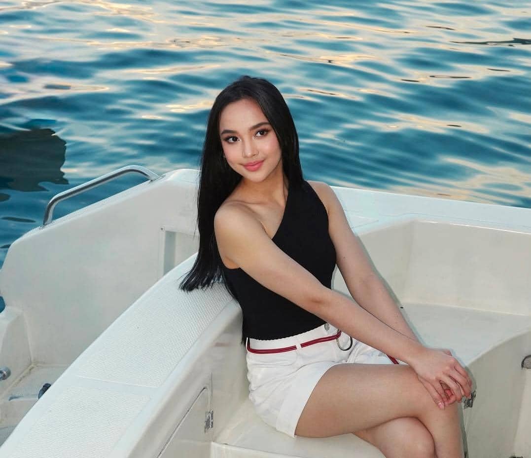 Seorang perempuan berambut panjang mengenakan atasan hitam tanpa lengan dan celana putih duduk di perahu dengan latar air biru tenang.