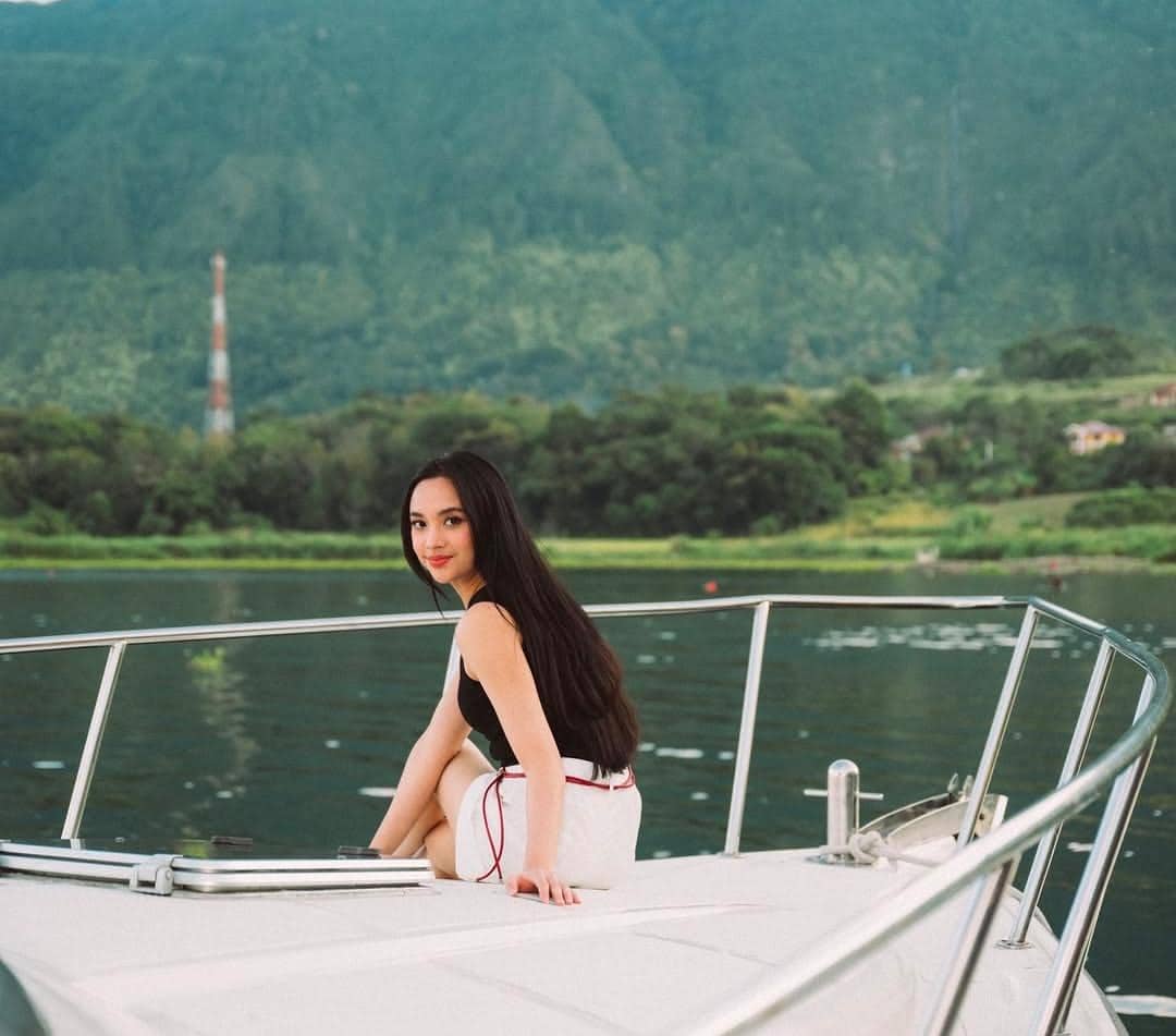 Seorang perempuan duduk di atas kapal menikmati pemandangan danau dan pegunungan hijau di Pulau Samosir yang menenangkan.
