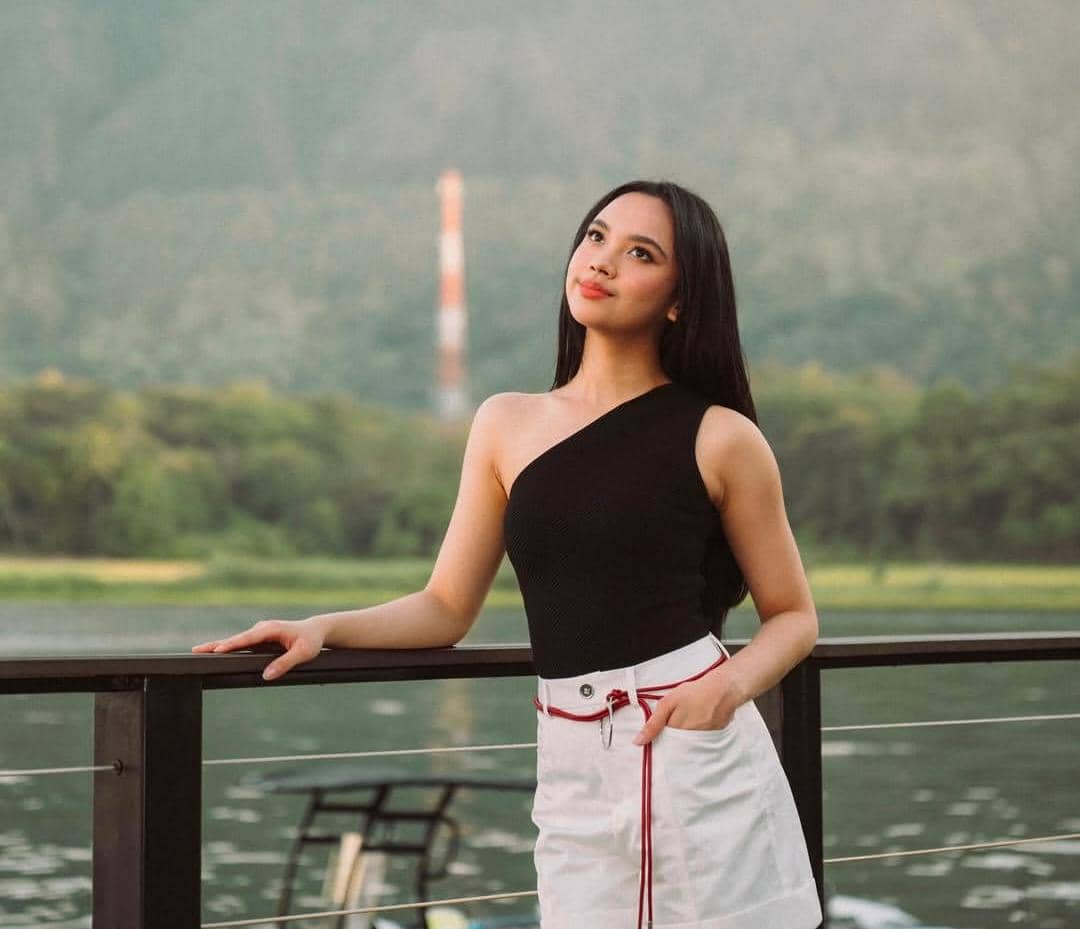 Seorang perempuan berambut panjang mengenakan atasan hitam tanpa lengan dan rok putih berpose di tepi danau dengan latar pegunungan hijau.
