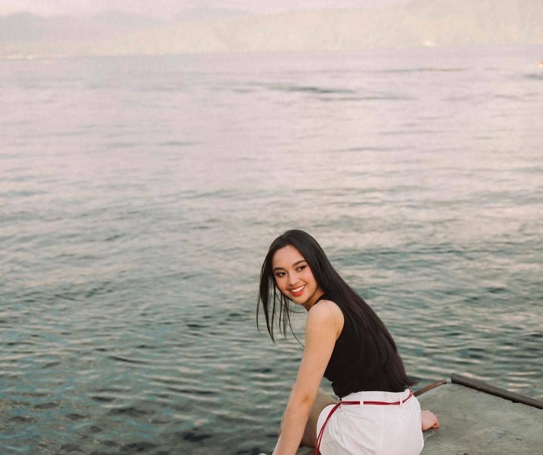 Seorang perempuan berambut panjang tersenyum sambil duduk di tepi danau dengan latar air jernih dan pegunungan di Pulau Samosir.