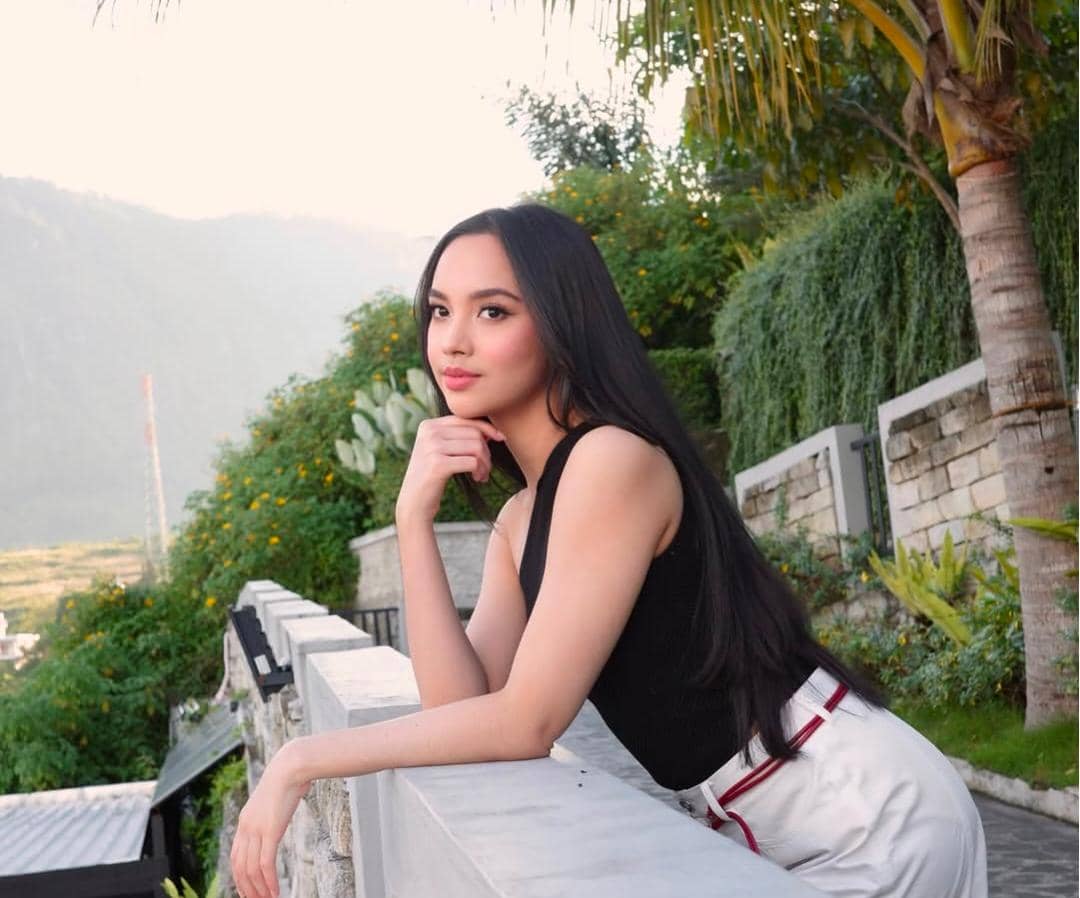 Seorang perempuan berambut panjang mengenakan atasan hitam dan celana putih berpose santai di area outdoor dengan latar pepohonan hijau.