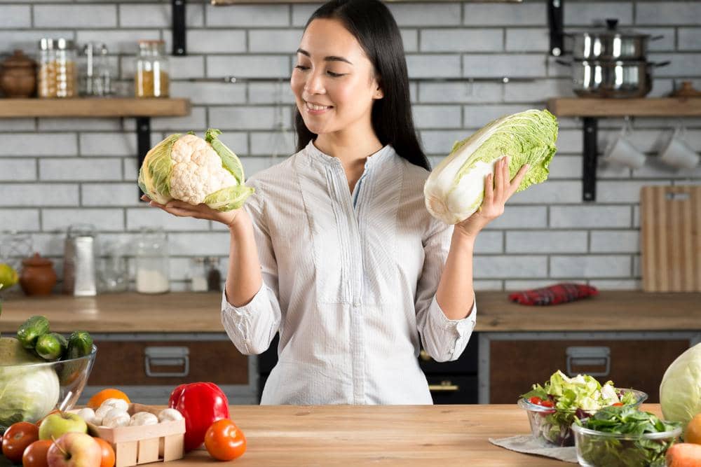 Seorang perempuan di dapur memegang kembang kol dan sawi putih sambil tersenyum, dengan berbagai sayuran segar di meja kayu.