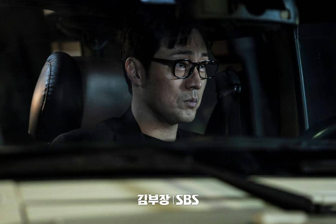 Seorang pria berkacamata duduk di dalam mobil pada malam hari dengan ekspresi serius dalam adegan drama Agent Kim Reactivated.