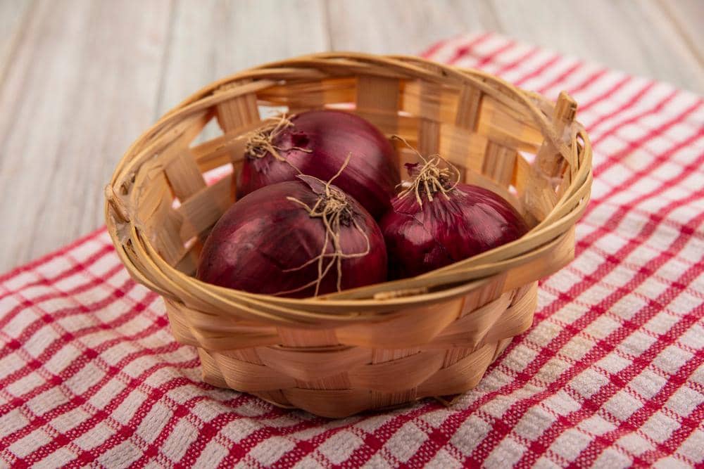 Beberapa bawang bombay merah segar di dalam keranjang anyaman bambu di atas kain bermotif kotak merah putih.