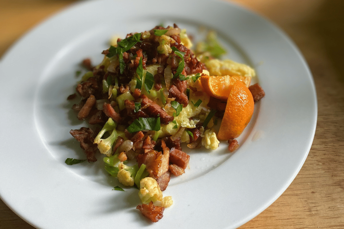 Salad orzo dengan potongan daging, sayuran hijau, dan irisan jeruk lemon disajikan di atas piring putih di atas meja kayu.