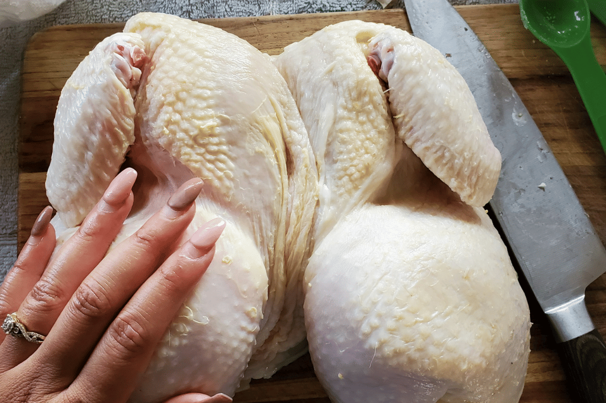 Tangan seseorang menyiapkan ayam utuh mentah di atas talenan kayu dengan pisau dapur dan sendok takar hijau di sampingnya.
