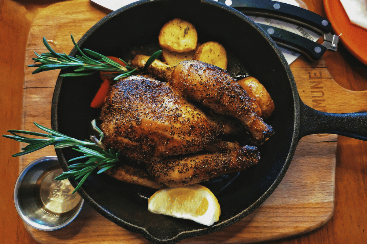 Ayam panggang berbumbu lada hitam disajikan dalam wajan besi dengan potongan kentang, irisan lemon, dan hiasan rosemary segar.
