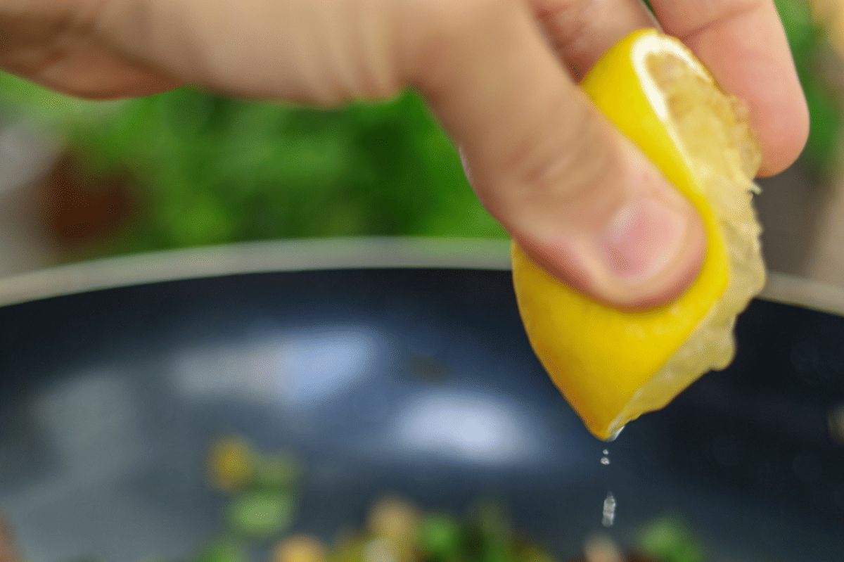 Tangan memeras potongan lemon segar di atas mangkuk berisi bahan salad orzo dengan rempah hijau di latar belakang.