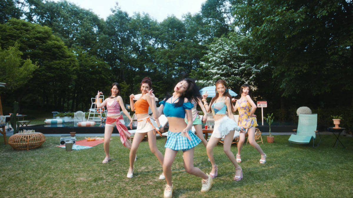 Para anggota BABYMONSTER menari dengan pakaian berwarna cerah di taman hijau dalam cuplikan video musik I LIKE IT.