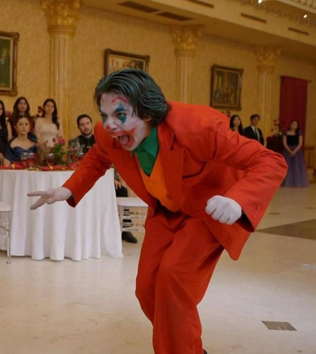 Seorang pria mengenakan kostum jas merah dan riasan wajah badut menirukan karakter Joker di sebuah ruangan pesta mewah.