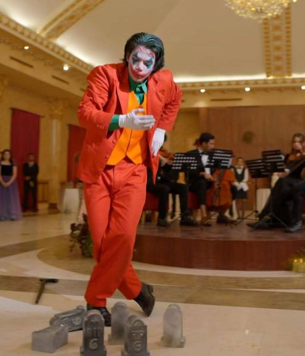 Seorang pria berdandan seperti badut dengan jas merah dan rompi oranye menari di aula mewah dengan orkestra di latar belakang.