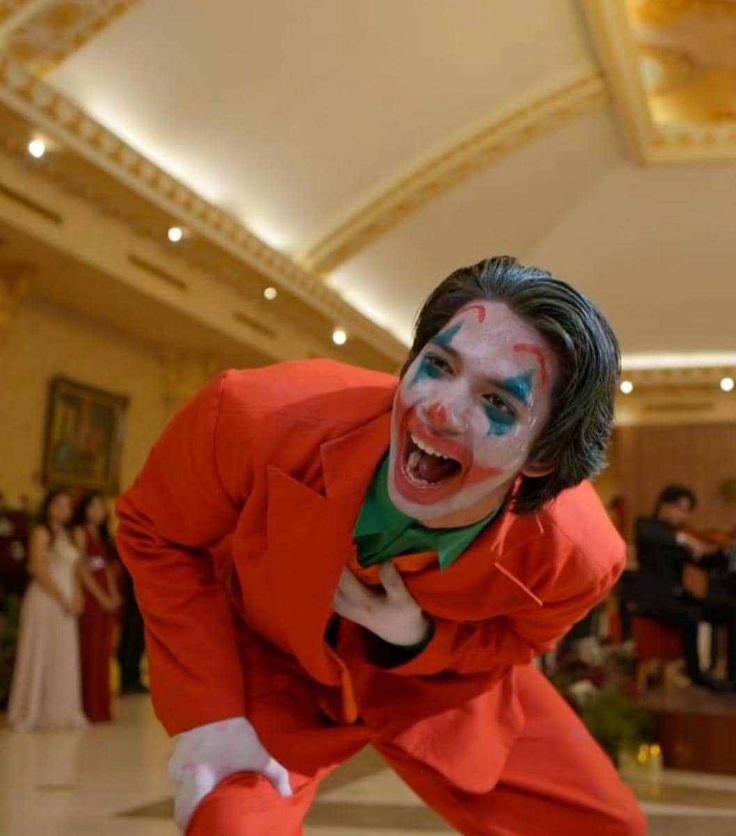 Seorang pria dengan riasan wajah badut berwarna merah, biru, dan putih mengenakan setelan jas merah sedang tertawa di ruangan mewah.