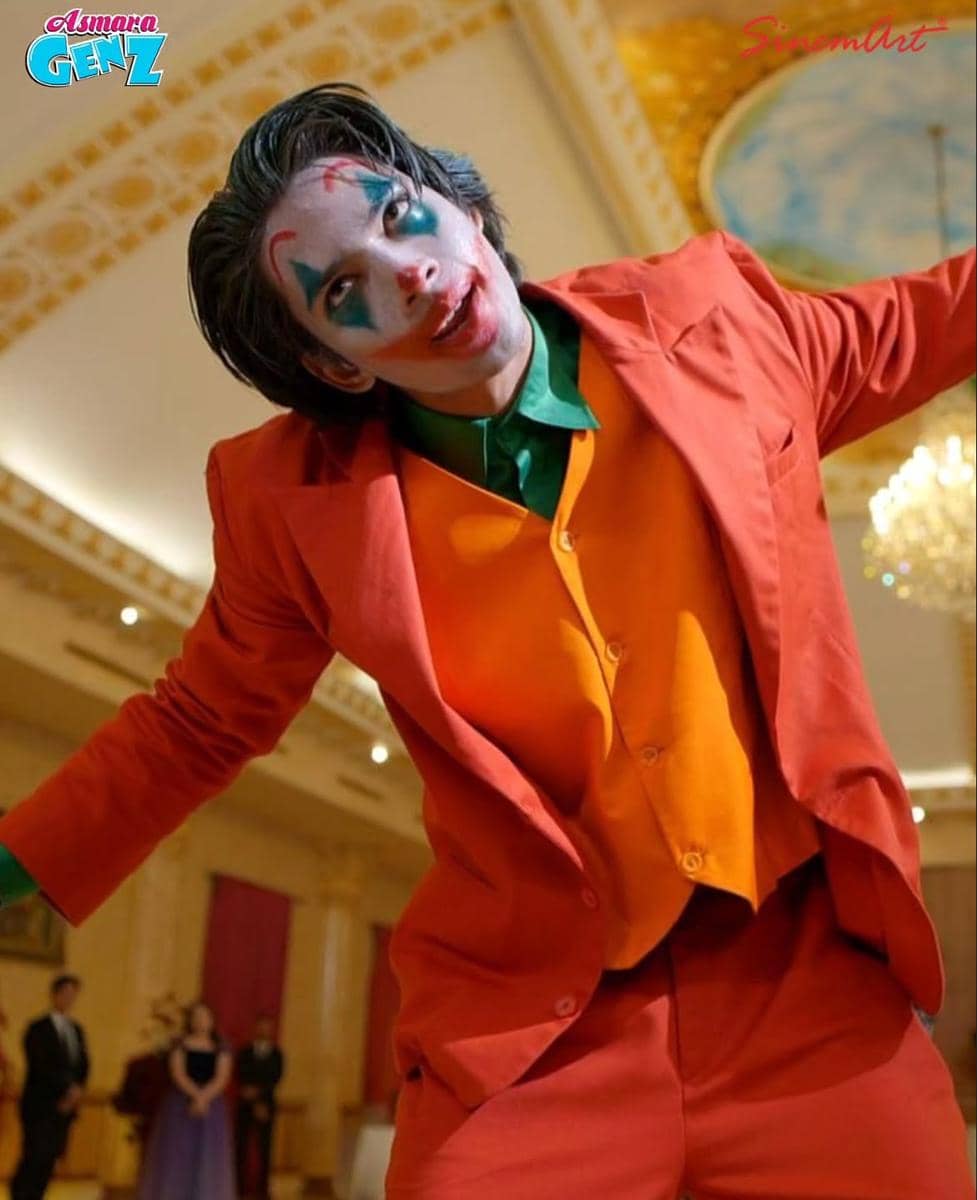 Seorang pria dengan riasan wajah badut bergaya Joker mengenakan setelan jas merah dan rompi oranye di ruangan mewah bercahaya hangat.