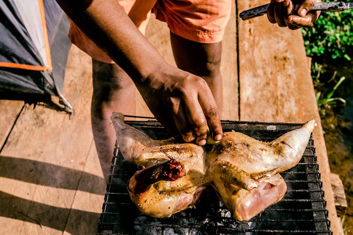Seseorang sedang memanggang ayam utuh di atas panggangan arang di luar ruangan dengan suasana cahaya matahari.
