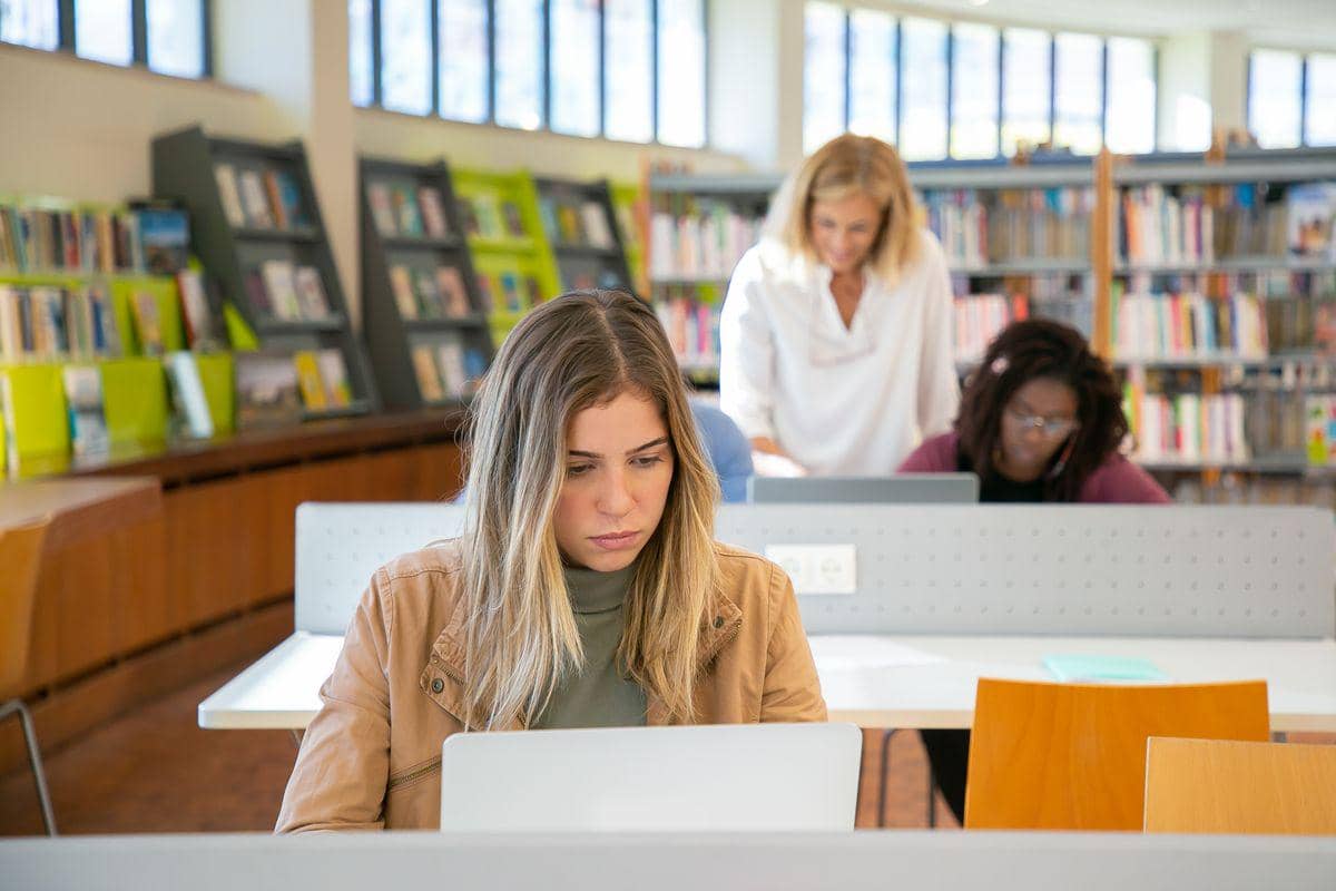 Beberapa orang bekerja menggunakan laptop di perpustakaan modern dengan rak buku di latar belakang dan suasana tenang.