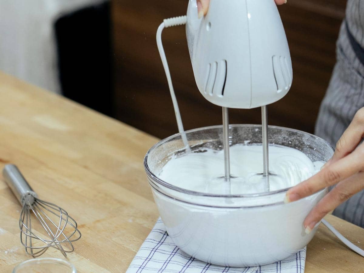 Seseorang sedang mengocok whipping cream dengan mixer listrik di dalam mangkuk kaca di atas meja dapur kayu.