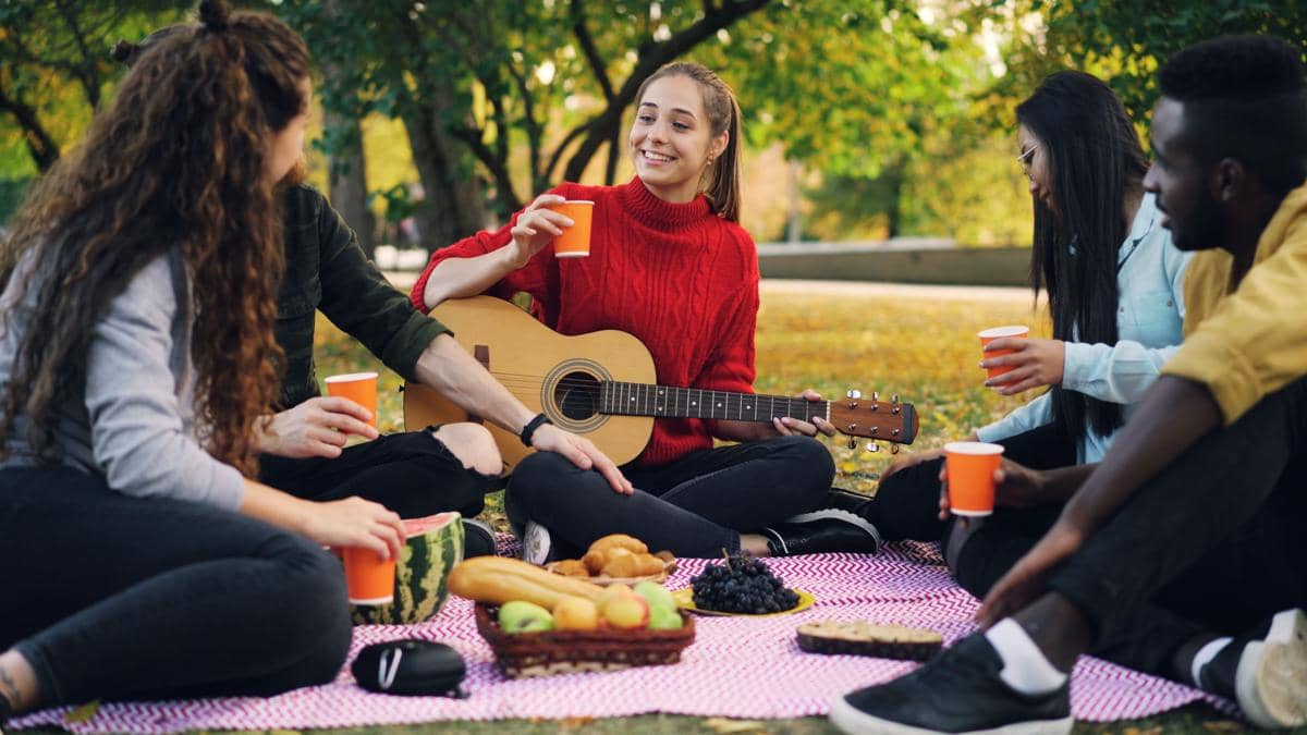 Sekelompok teman duduk di taman sambil piknik, menikmati minuman dan makanan, salah satu memainkan gitar dengan suasana santai.