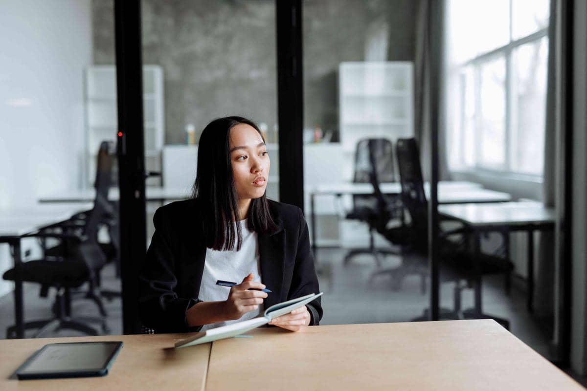 Seorang wanita duduk di meja kantor dengan buku catatan dan pena di tangan, menatap ke arah jendela dengan ekspresi berpikir.