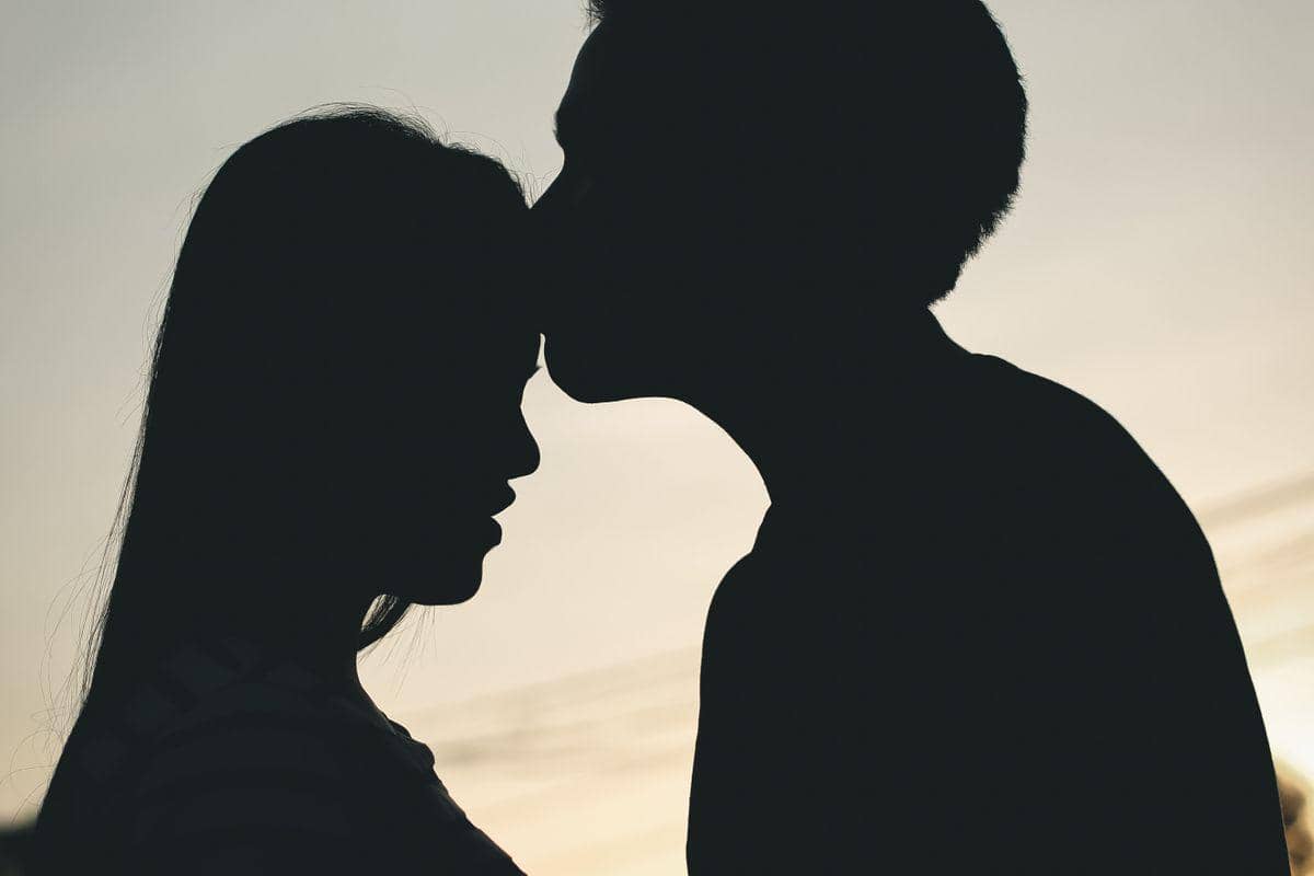 Siluet seorang pria mencium kening seorang wanita di bawah cahaya senja yang lembut, menciptakan suasana hangat dan penuh kasih.