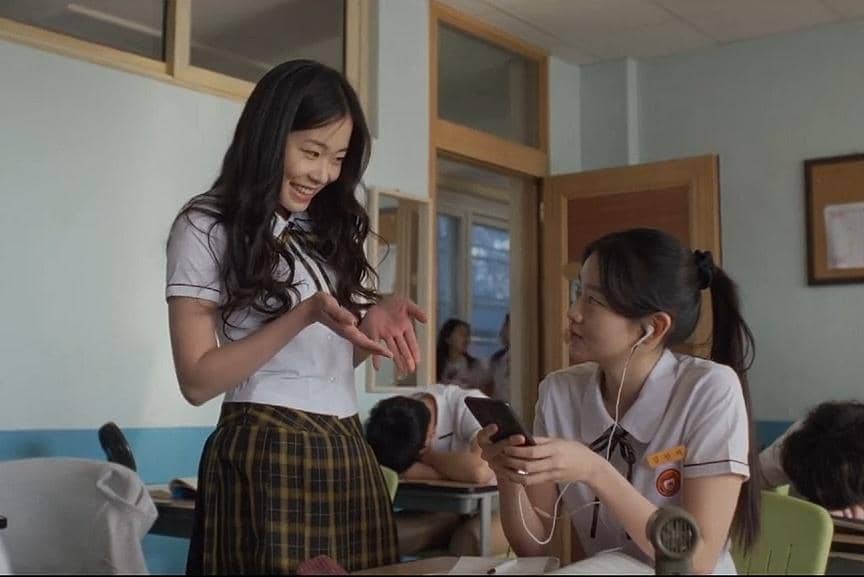 Dua siswi berseragam sekolah sedang berbincang di ruang kelas, satu berdiri sambil tersenyum dan satu duduk memegang ponsel.