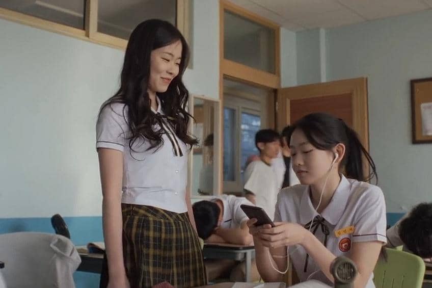 Dua siswi berseragam sekolah sedang berbincang di dalam kelas, salah satunya memegang ponsel sambil mengenakan earphone.