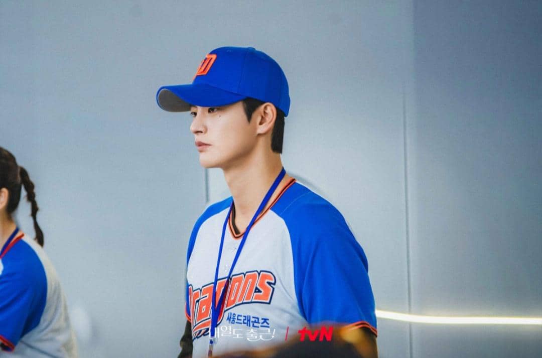 Seorang pria mengenakan seragam bisbol biru putih dan topi biru dengan logo oranye dalam cuplikan drama Korea See You at Work Tomorrow.