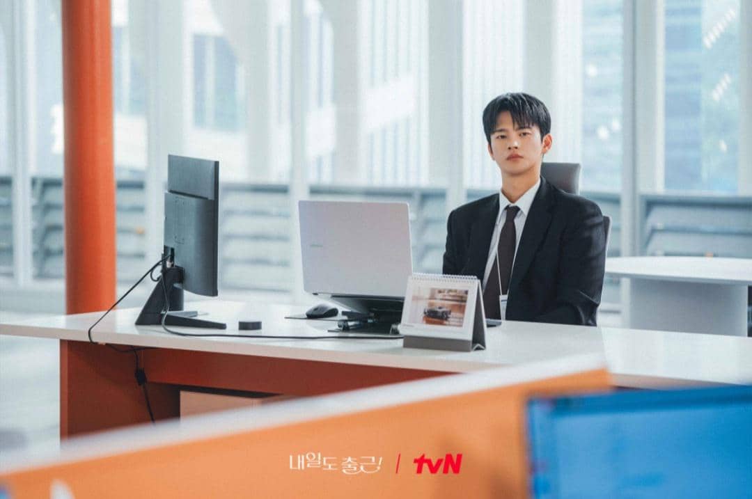 Seorang pria mengenakan setelan jas hitam duduk di meja kantor modern dengan laptop dan monitor di depannya dalam cuplikan drama See You at Work Tomorrow.
