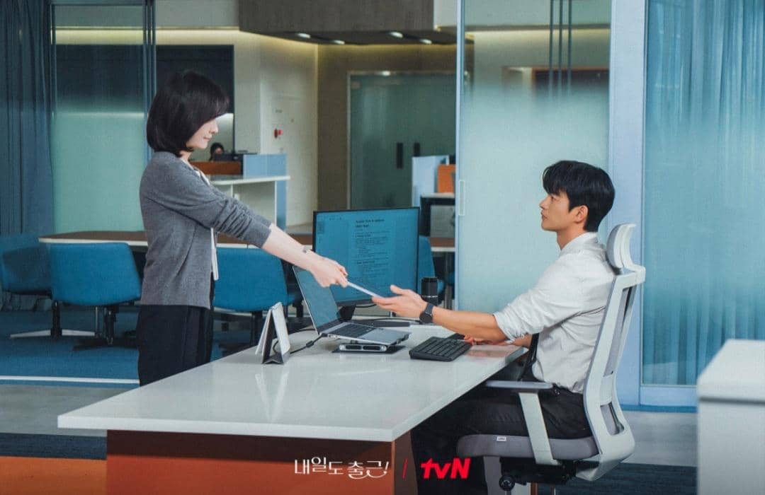 Dua karyawan sedang berinteraksi di kantor modern, seorang wanita menyerahkan dokumen kepada pria yang duduk di depan komputer.
