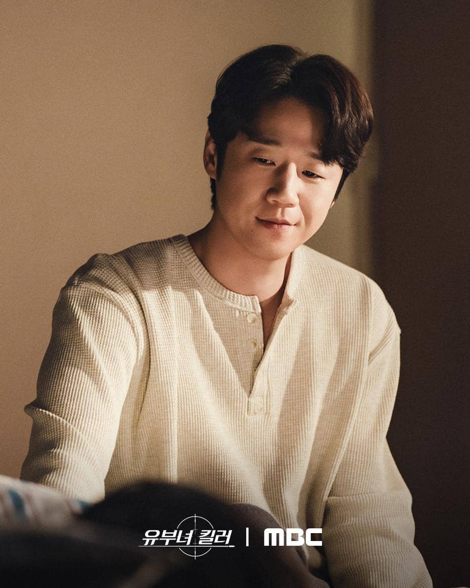 Seorang pria mengenakan sweater krem tersenyum lembut di ruangan bercahaya hangat dalam adegan drama A Bona Fide Killer produksi MBC.