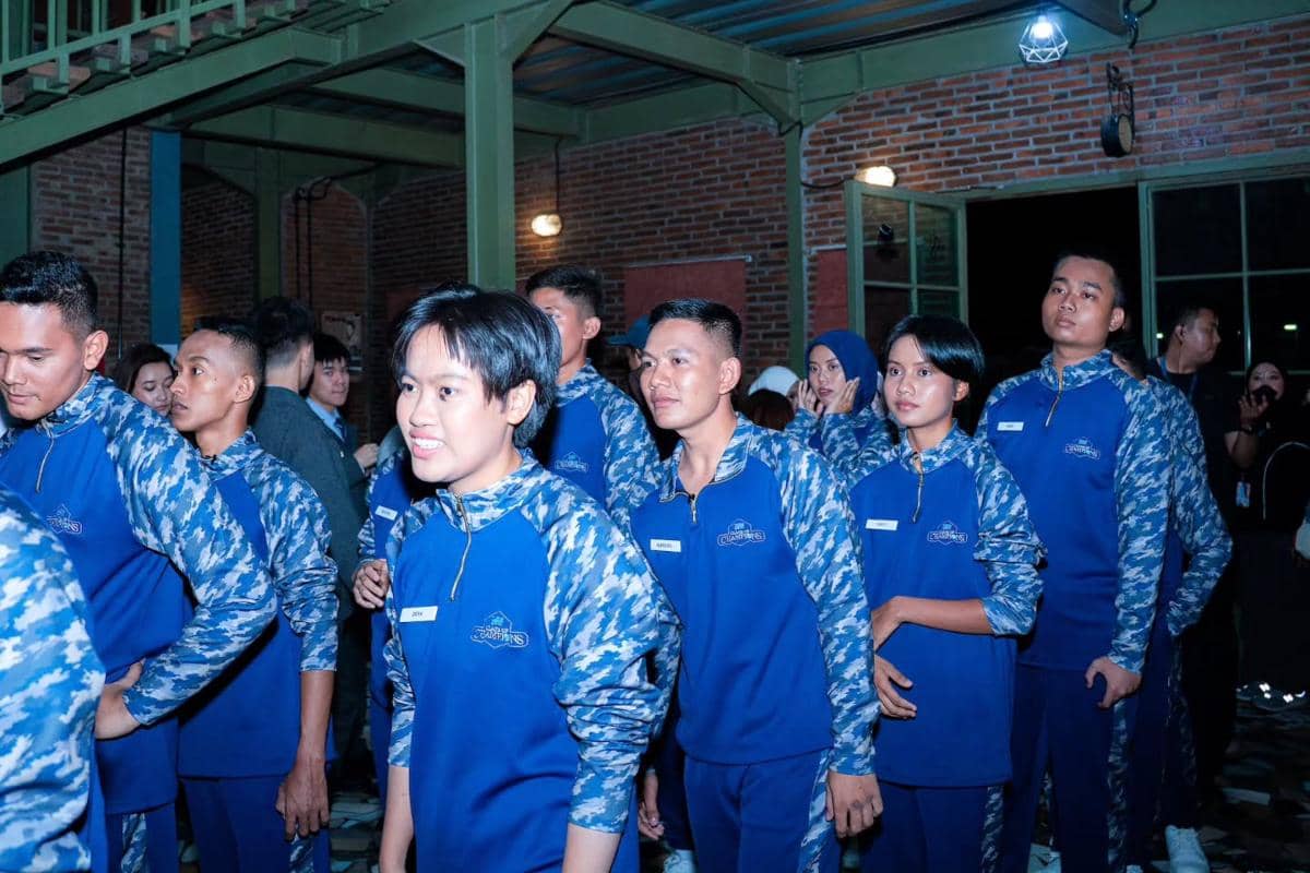 Para peserta Clash of Champions mengenakan seragam biru bermotif kamuflase berdiri berbaris di sebuah ruangan dengan dinding bata dan pencahayaan malam.