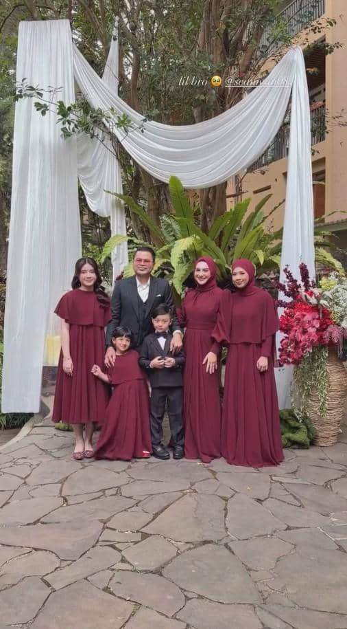 Sebuah keluarga berpose di acara pernikahan dengan latar dekorasi kain putih dan tanaman hijau, mengenakan busana formal bernuansa marun.