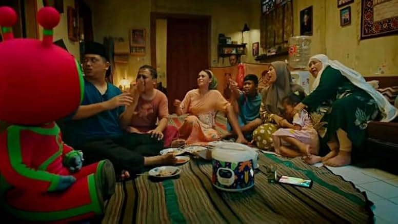 Sekelompok orang duduk di ruang tamu sederhana bersama karakter berkostum merah hijau berbentuk alien dalam adegan film Foufo.