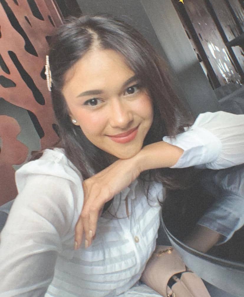 Seorang perempuan berambut panjang mengenakan blus putih tersenyum sambil bersandar di meja dengan pencahayaan lembut di ruangan modern.