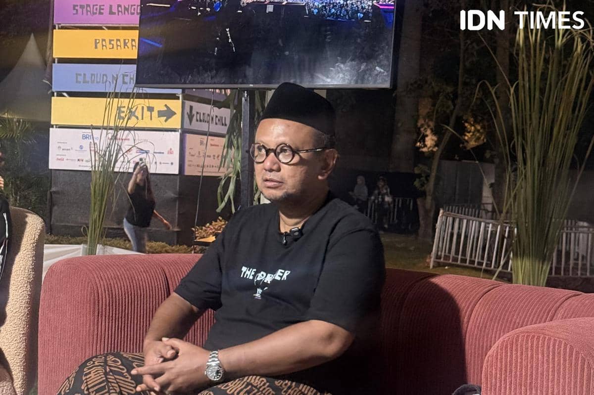 Seorang pria mengenakan kemeja hitam dan peci duduk di sofa merah saat wawancara di area Prambanan Jazz Festival 2026 pada malam hari.