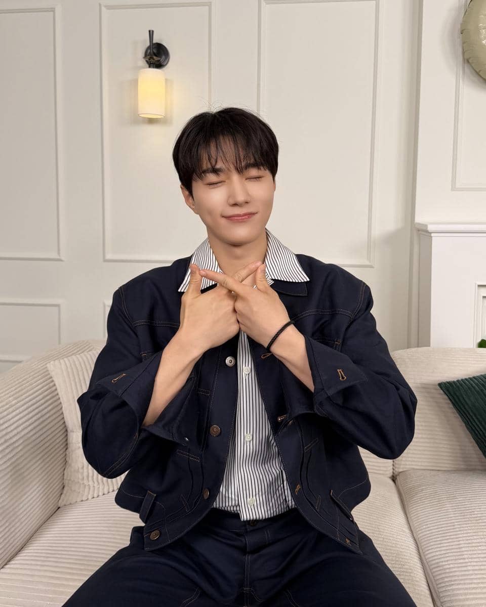 Seorang pria berambut hitam mengenakan jaket denim biru tua dan kemeja bergaris duduk di sofa dengan mata tertutup sambil tersenyum.