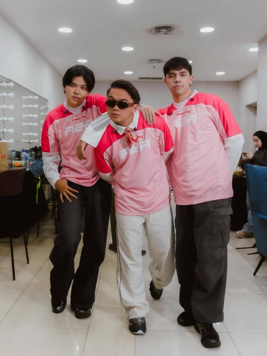 Tiga pria mengenakan kaus berwarna merah muda dengan logo Good Day berpose santai di ruang ganti yang terang dan modern.
