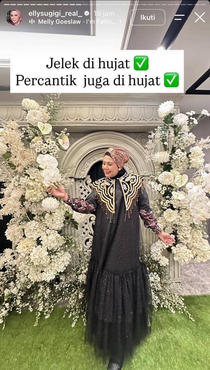 Seorang wanita berhijab mengenakan gaun hitam berhias detail emas berpose di depan dekorasi bunga putih yang mewah dan elegan.