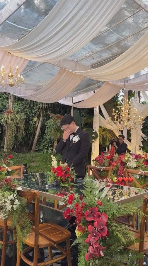 Seorang pria mengenakan setelan jas hitam dengan bunga di dada berdiri di dekorasi pernikahan bertema taman dengan hiasan bunga merah dan putih.