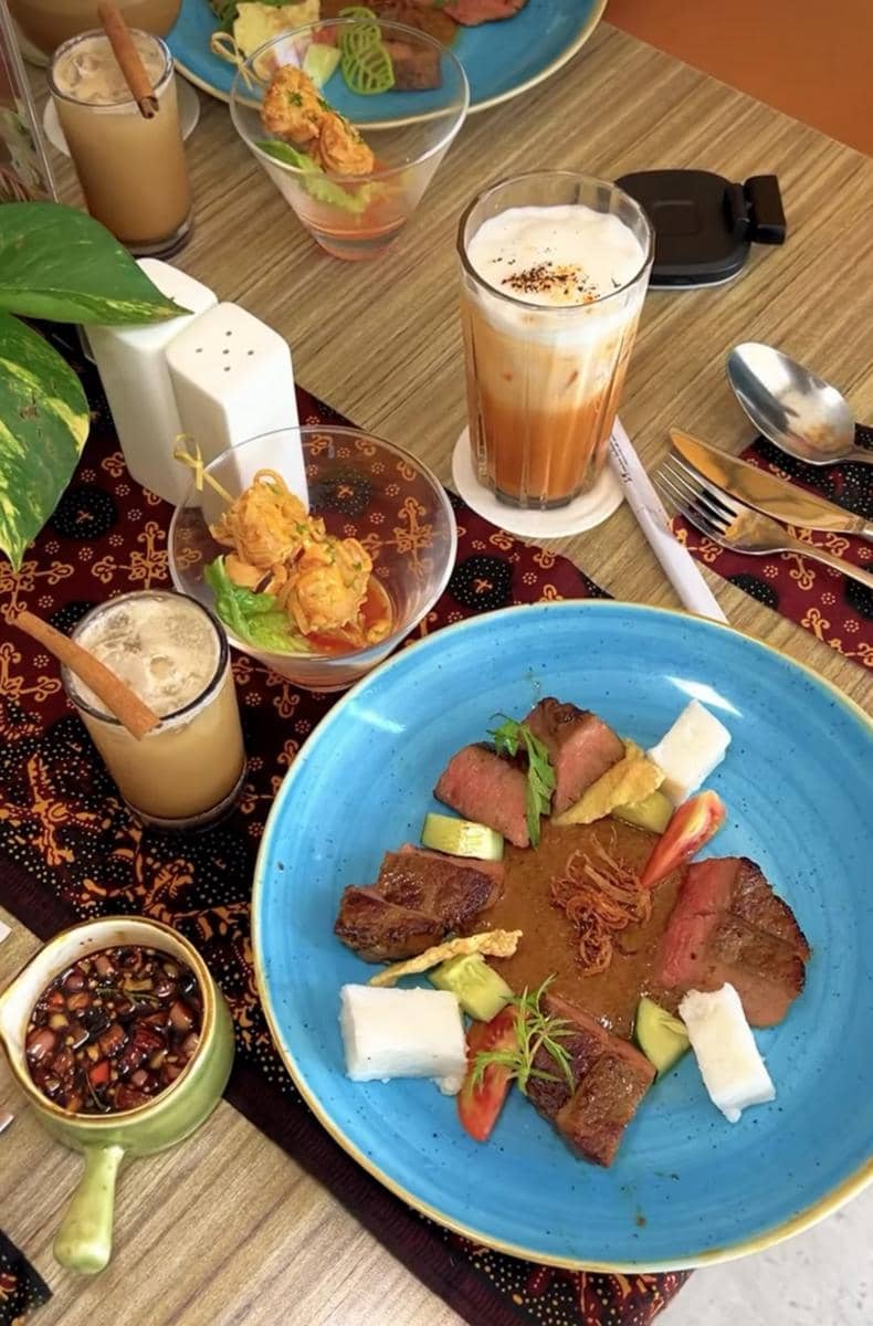 Hidangan daging dengan lontong, sayuran, dan saus kacang disajikan di piring biru bersama minuman kopi dan teh di meja restoran.