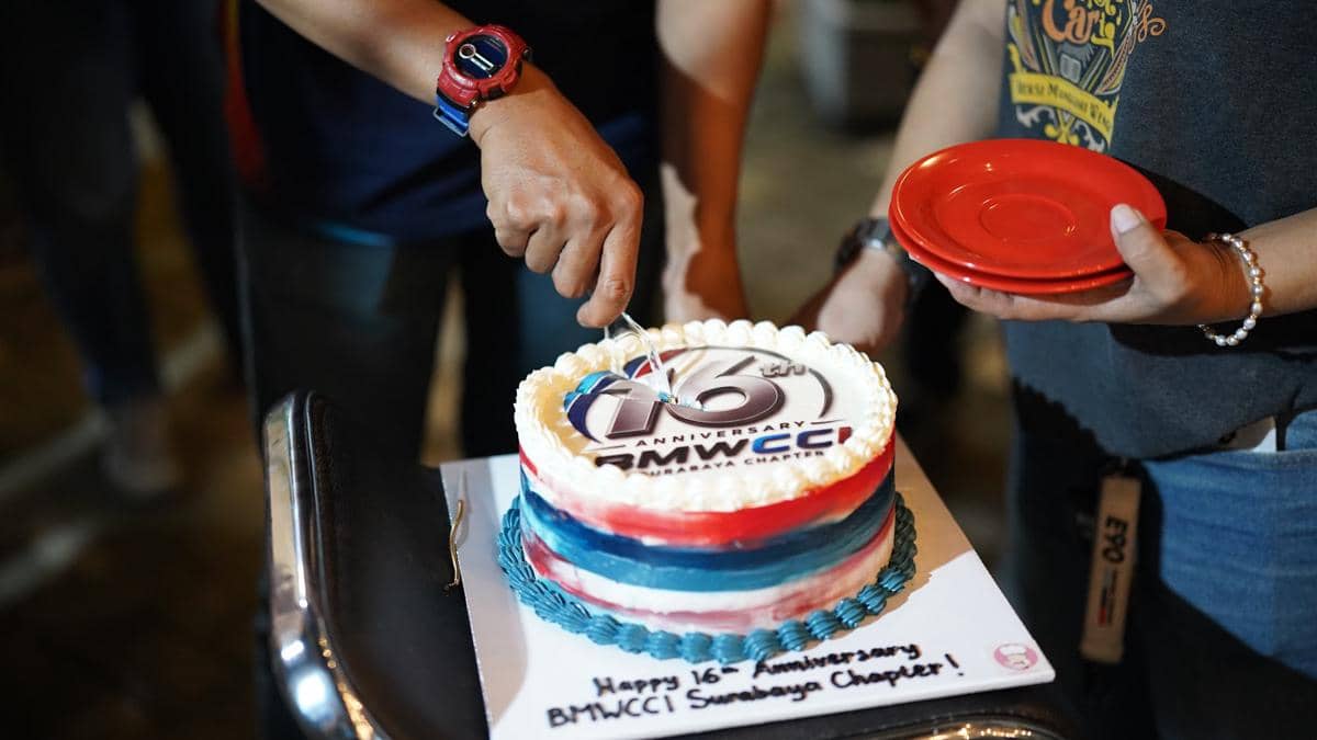 Kue ulang tahun bertema 16th Anniversary BMWCCI dengan hiasan warna merah, biru, dan putih sedang dipotong oleh dua orang.