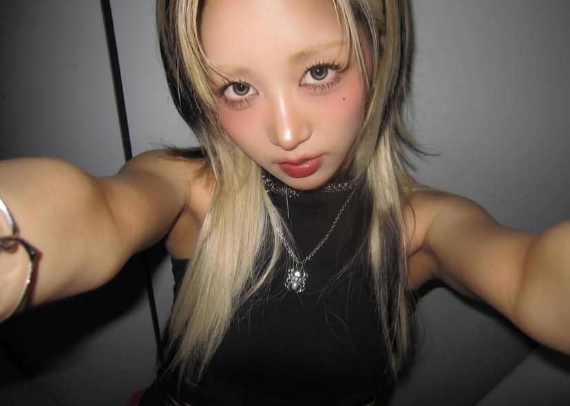 Seorang perempuan berambut pirang dengan riasan tegas dan mengenakan atasan hitam berpose selfie di ruangan bercahaya redup.