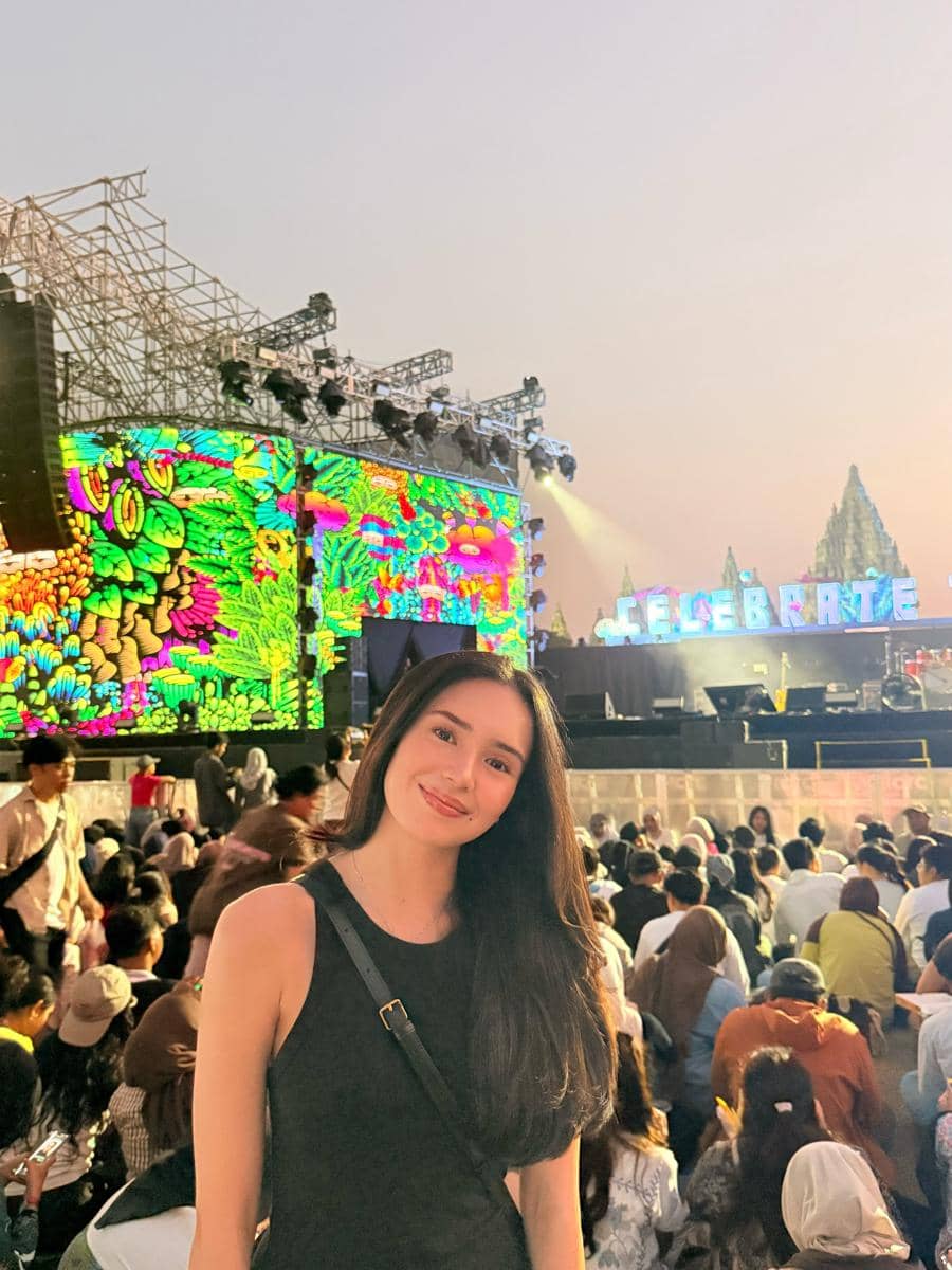 Seorang perempuan berpose di depan panggung berwarna-warni di Prambanan Jazz Festival 2026 dengan latar penonton yang ramai.
