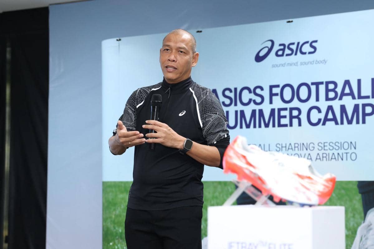 Seorang pembicara mengenakan pakaian olahraga hitam berbicara di acara ASICS Football Summer Camp 2026 dengan latar belakang spanduk dan sepatu baru di depan.