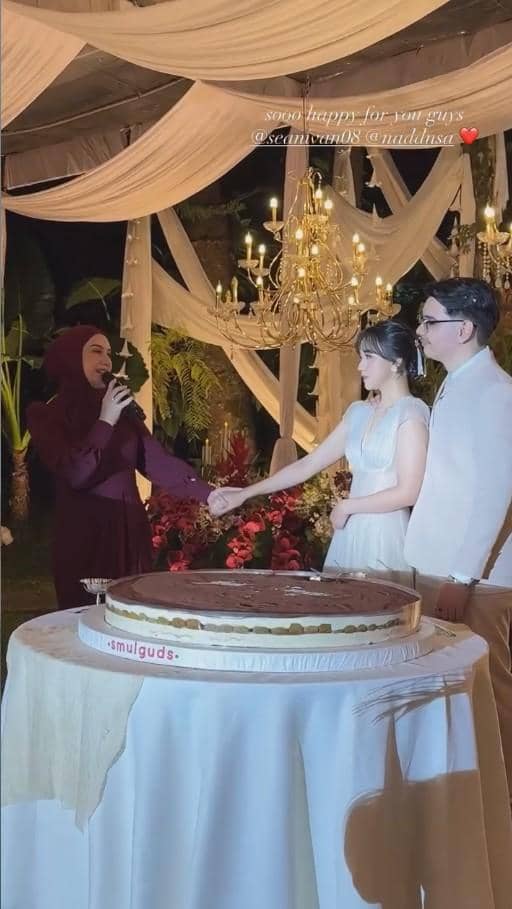 Pasangan pengantin berdiri di depan kue besar sambil bergandengan tangan di pesta pernikahan dengan dekorasi lampu gantung elegan.