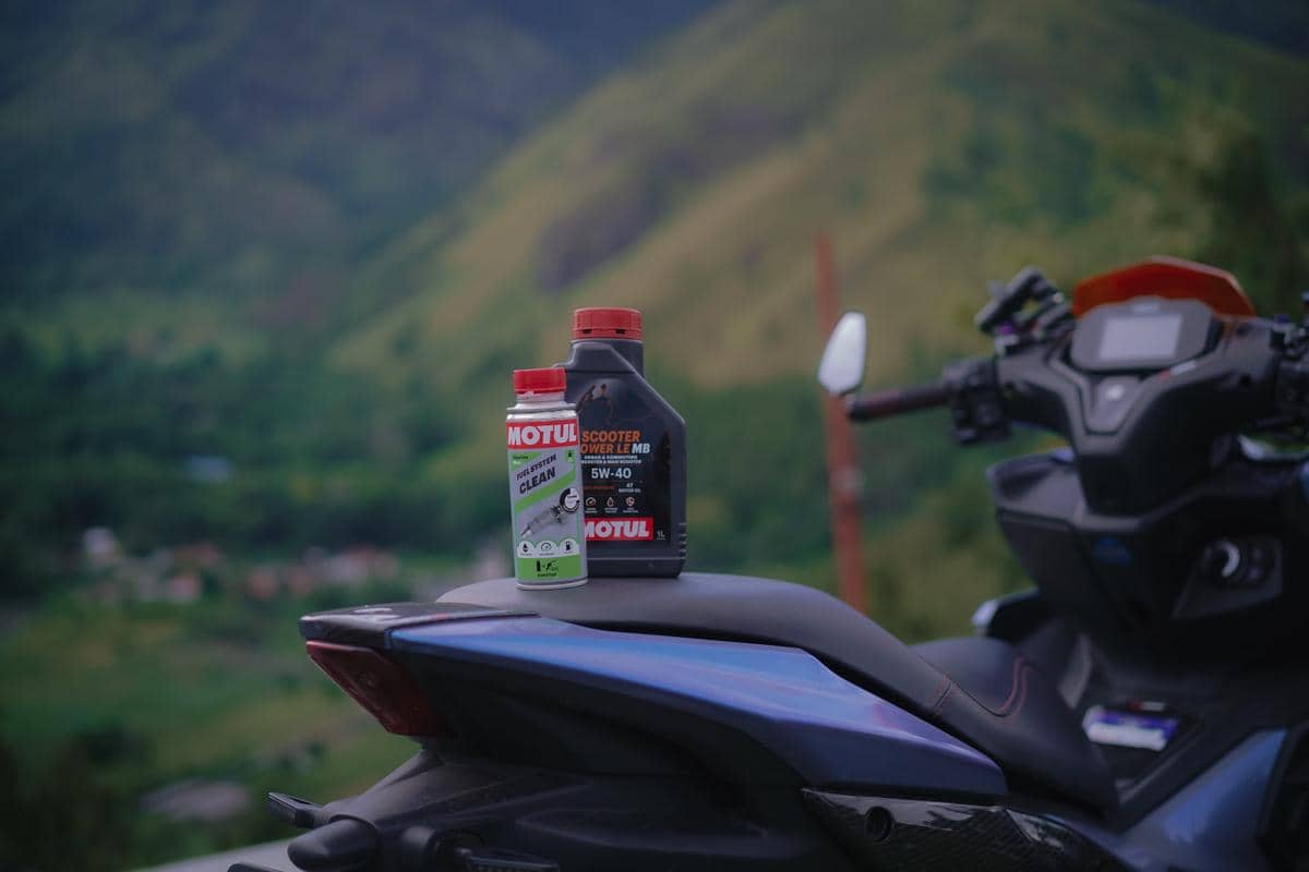 Dua botol pelumas Motul diletakkan di jok motor dengan latar belakang pemandangan pegunungan hijau yang indah.