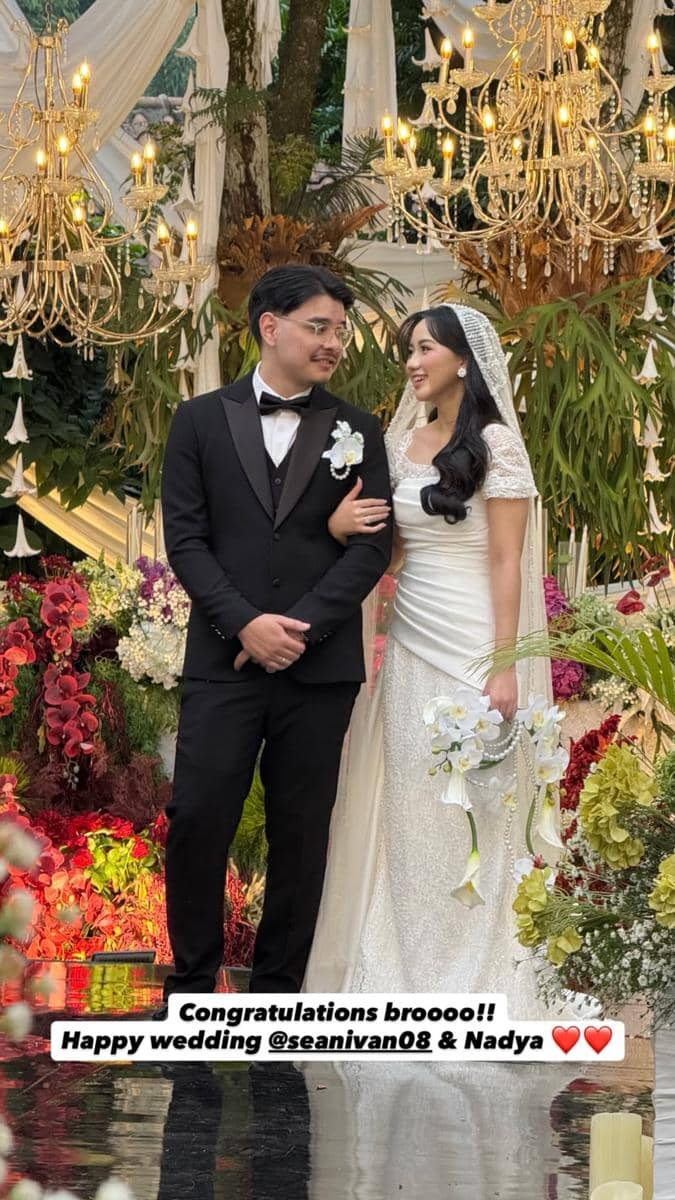 Pasangan pengantin berdiri saling menatap di pelaminan berhias bunga dan lampu gantung mewah, mengenakan gaun putih dan setelan jas hitam.
