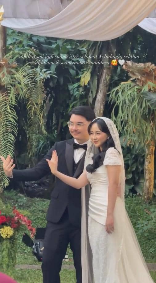 Pasangan pengantin berpose bersama di taman saat pernikahan, sang mempelai pria mengenakan setelan jas hitam dan mempelai wanita memakai gaun putih berhias kerudung.
